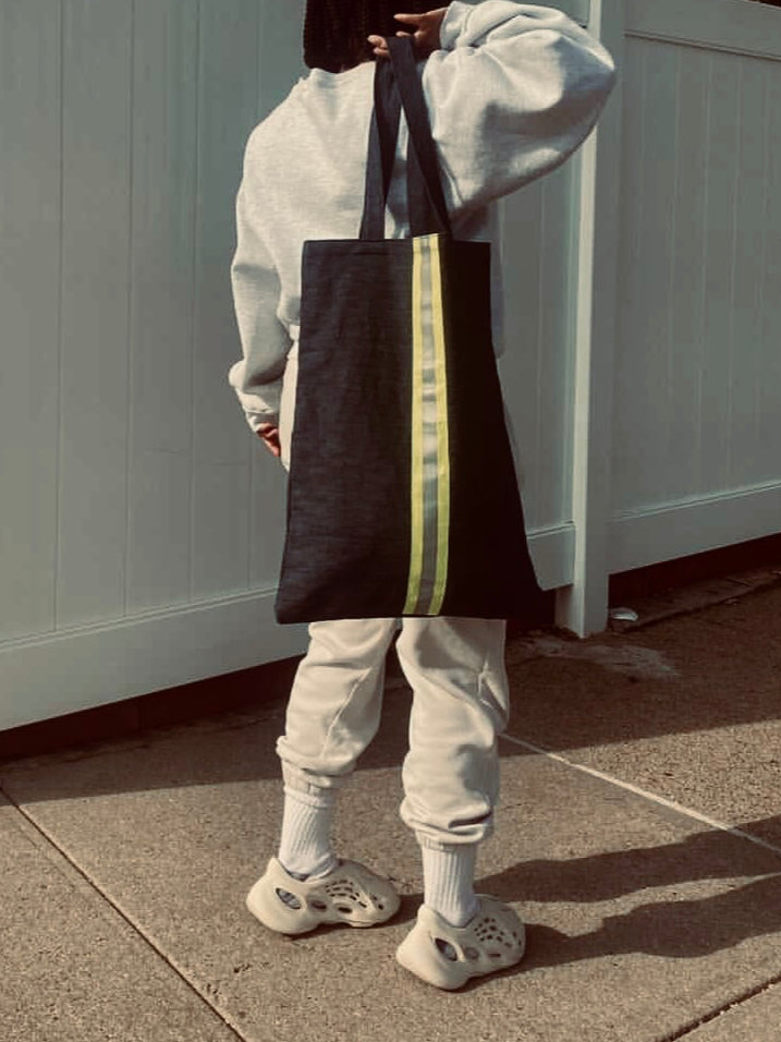 Person holding a black tote bag with yellow stripes against a white wall.